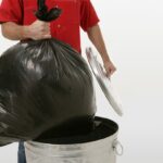 The torso of a man in a red shirt taking out a black bag of trash