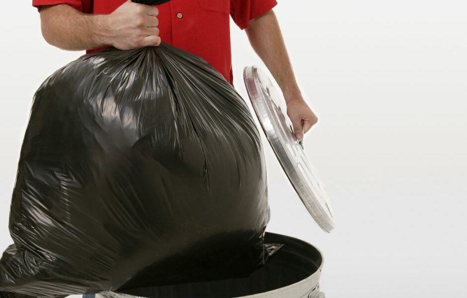 The torso of a man in a red shirt taking out a black bag of trash